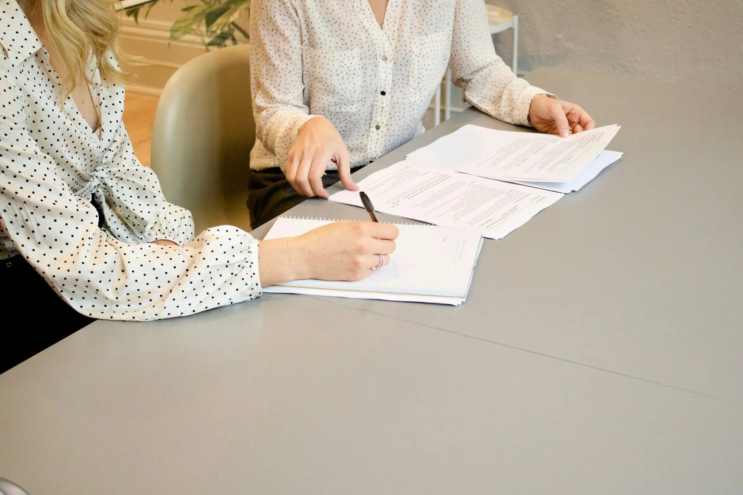 Dos mujeres revisando documentos en una mesa gris; una escribe en un cuaderno mientras la otra señala unas hojas impresas